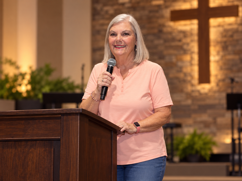Chris speaking with a microphone at a church podium, a wooden cross behind her
